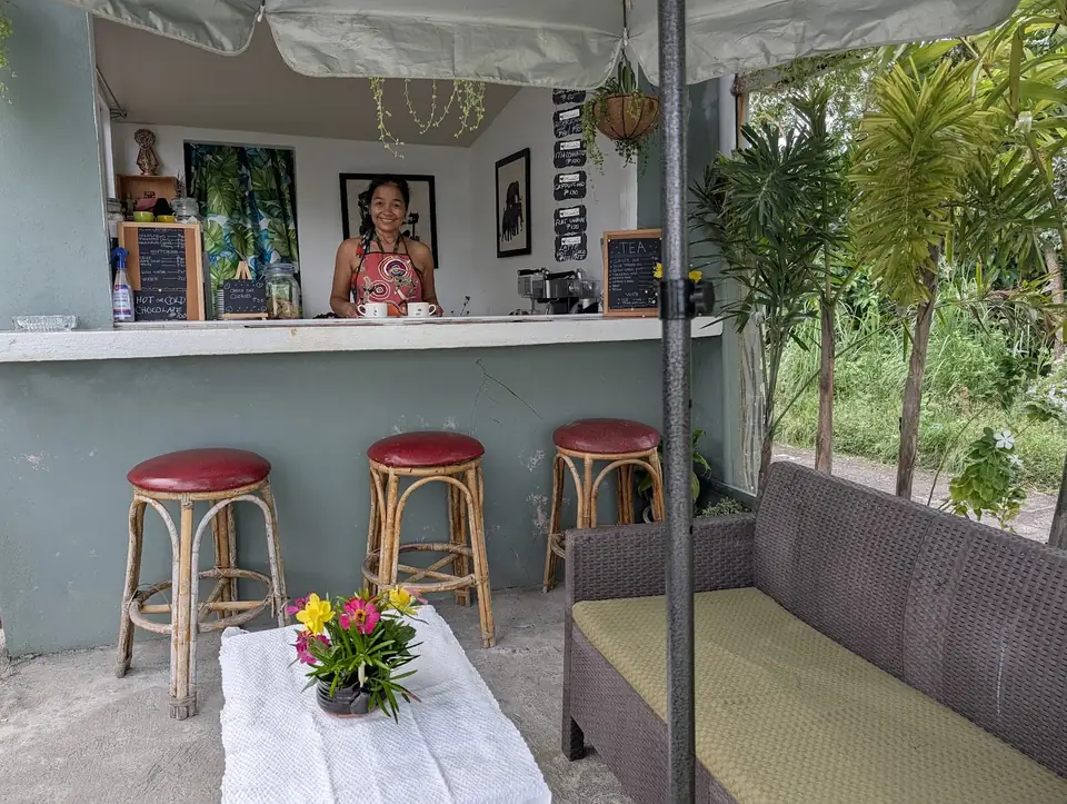 Ang nakangiting may-ari ng Kape Parola sa likod ng bukas na counter, kasama ang mga rattan stool at upuan sa hardin